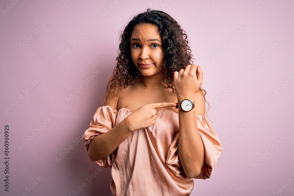 Young beautiful woman with curly hair wearing casual t-shirt standing over pink background In hurry pointing to watch time, impatience, looking at the camera with relaxed expression Young beautiful woman with curly hair wearing casual t-shirt standing over pink background In hurry pointing to watch time, impatience, looking at the camera with relaxed expression