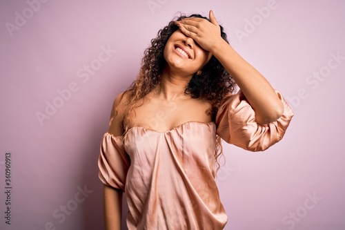 Young beautiful woman with curly hair wearing casual t-shirt standing over pink background smiling and laughing with hand on face covering eyes for surprise. Blind concept.