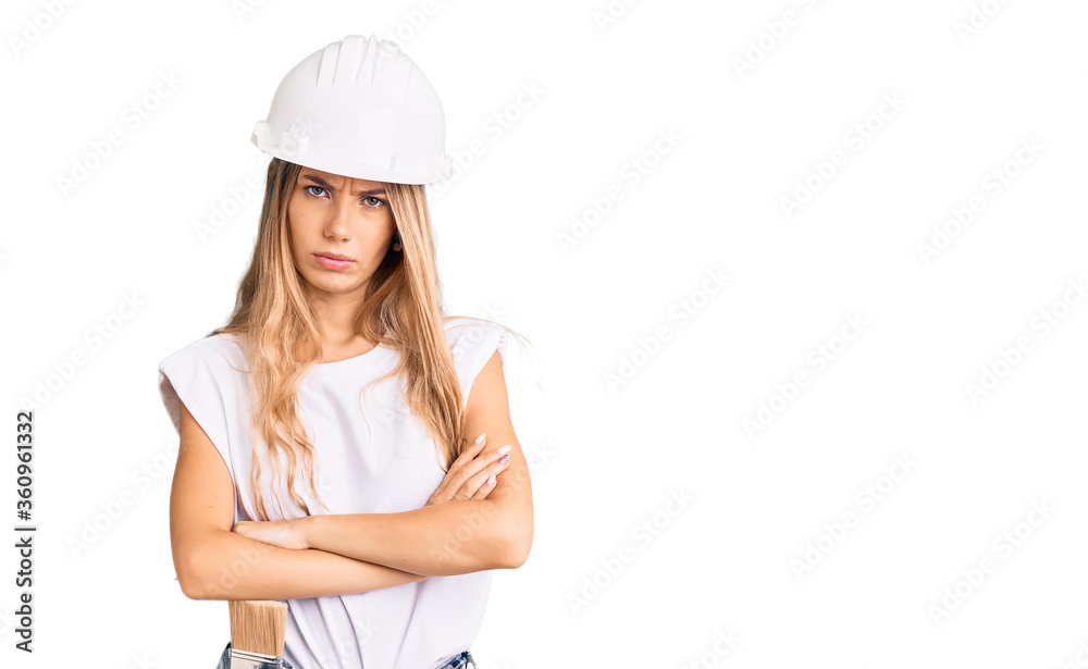 Beautiful caucasian woman with blonde hair wearing hardhat and painter clothes skeptic and nervous, disapproving expression on face with crossed arms. negative person.