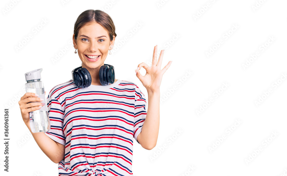 Beautiful caucasian woman with blonde hair wearing sportswear and headphones drinking a bottle of water doing ok sign with fingers, smiling friendly gesturing excellent symbol Beautiful caucasian woman with blonde hair wearing sportswear and headphones drinking a bottle of water doing ok sign with fingers, smiling friendly gesturing excellent symbol