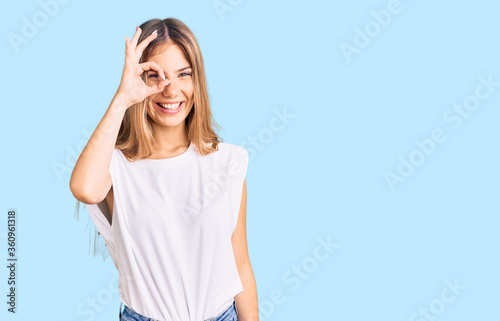 Beautiful caucasian woman with blonde hair wearing casual white tshirt doing ok gesture with hand smiling, eye looking through fingers with happy face.