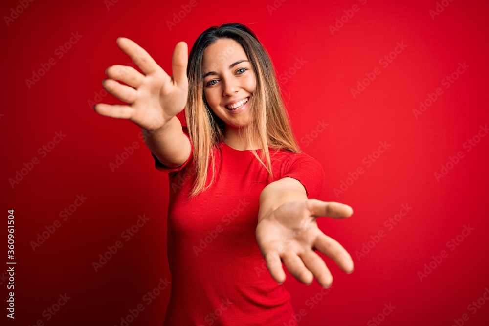 Young beautiful blonde woman with blue eyes wearing casual t-shirt over red background looking at the camera smiling with open arms for hug. Cheerful expression embracing happiness. Young beautiful blonde woman with blue eyes wearing casual t-shirt over red background looking at the camera smiling with open arms for hug. Cheerful expression embracing happiness.