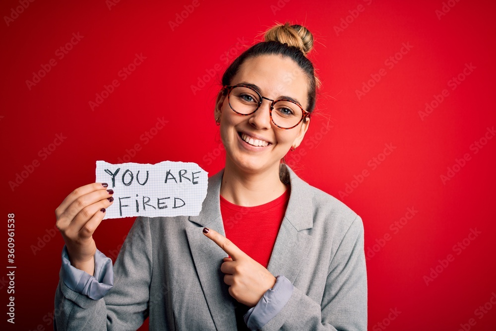 Young beautiful blonde woman with blue eyes holding paper with you are fired message very happy pointing with hand and finger Young beautiful blonde woman with blue eyes holding paper with you are fired message very happy pointing with hand and finger