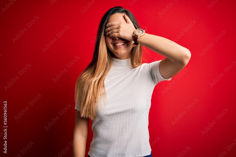 Beautiful blonde woman with blue eyes wearing casual white t-shirt over red background smiling and laughing with hand on face covering eyes for surprise. Blind concept. Beautiful blonde woman with blue eyes wearing casual white t-shirt over red background smiling and laughing with hand on face covering eyes for surprise. Blind concept.