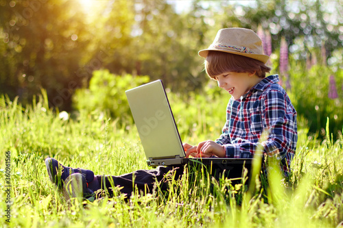Kid with a laptop outdoors in the summer. Сhild in a park on distance learning. Online communication with relatives and friends. Video call of a grandson to grandparents. Summertime positive scene.