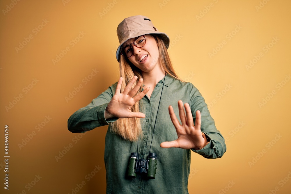 Beautiful blonde explorer woman with blue eyes wearing hat and glasses using binoculars afraid and terrified with fear expression stop gesture with hands, shouting in shock. Panic concept. Beautiful blonde explorer woman with blue eyes wearing hat and glasses using binoculars afraid and terrified with fear expression stop gesture with hands, shouting in shock. Panic concept.