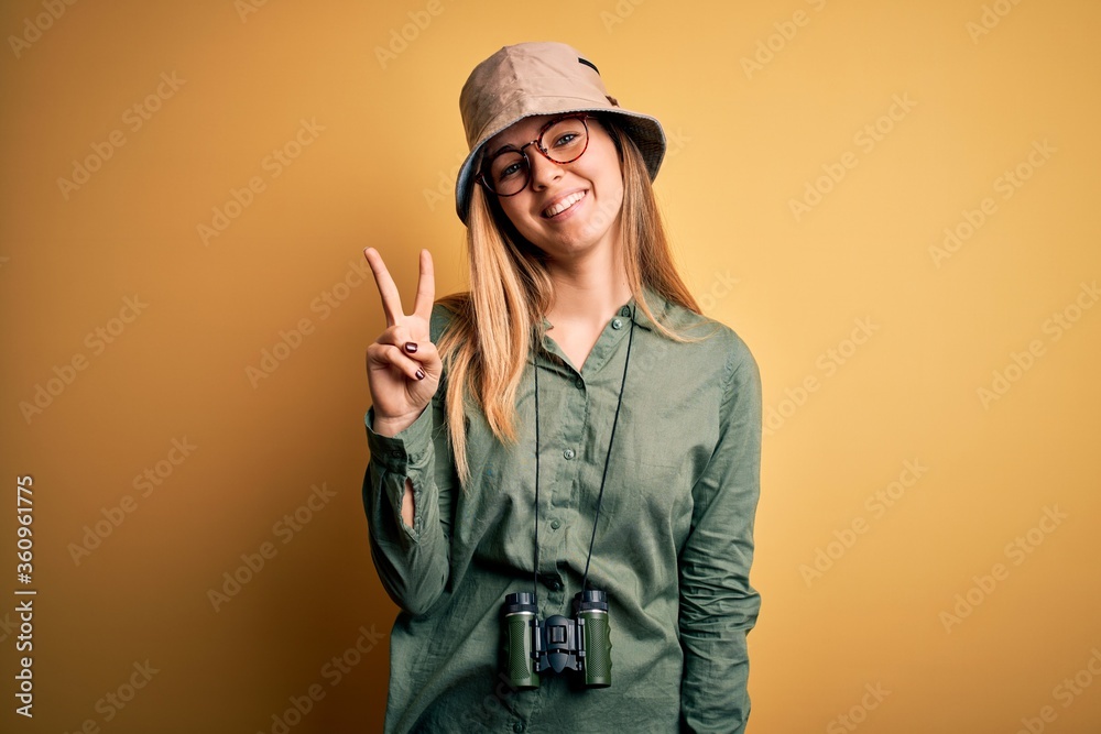 Beautiful blonde explorer woman with blue eyes wearing hat and glasses using binoculars showing and pointing up with fingers number two while smiling confident and happy. Beautiful blonde explorer woman with blue eyes wearing hat and glasses using binoculars showing and pointing up with fingers number two while smiling confident and happy.