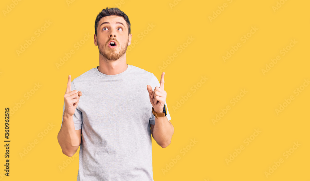 Handsome young man with bear wearing casual tshirt amazed and surprised looking up and pointing with fingers and raised arms. Handsome young man with bear wearing casual tshirt amazed and surprised looking up and pointing with fingers and raised arms.