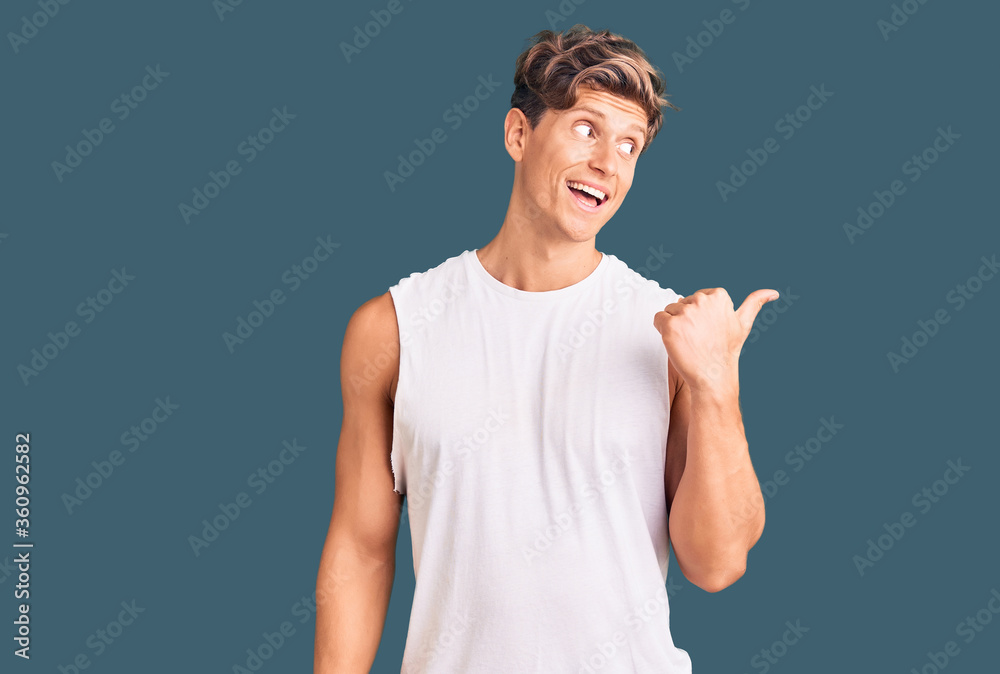 Young handsome man wearing sleveeless t shirt smiling with happy face looking and pointing to the side with thumb up. Young handsome man wearing sleveeless t shirt smiling with happy face looking and pointing to the side with thumb up.