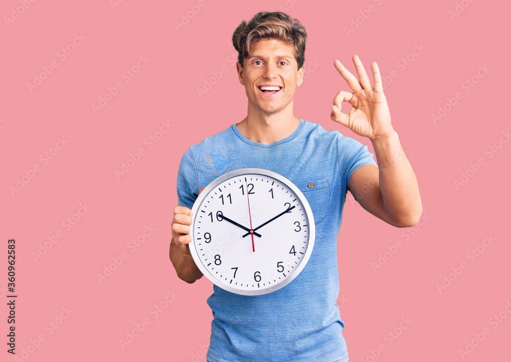 Young handsome man holding big clock doing ok sign with fingers, smiling friendly gesturing excellent symbol Young handsome man holding big clock doing ok sign with fingers, smiling friendly gesturing excellent symbol