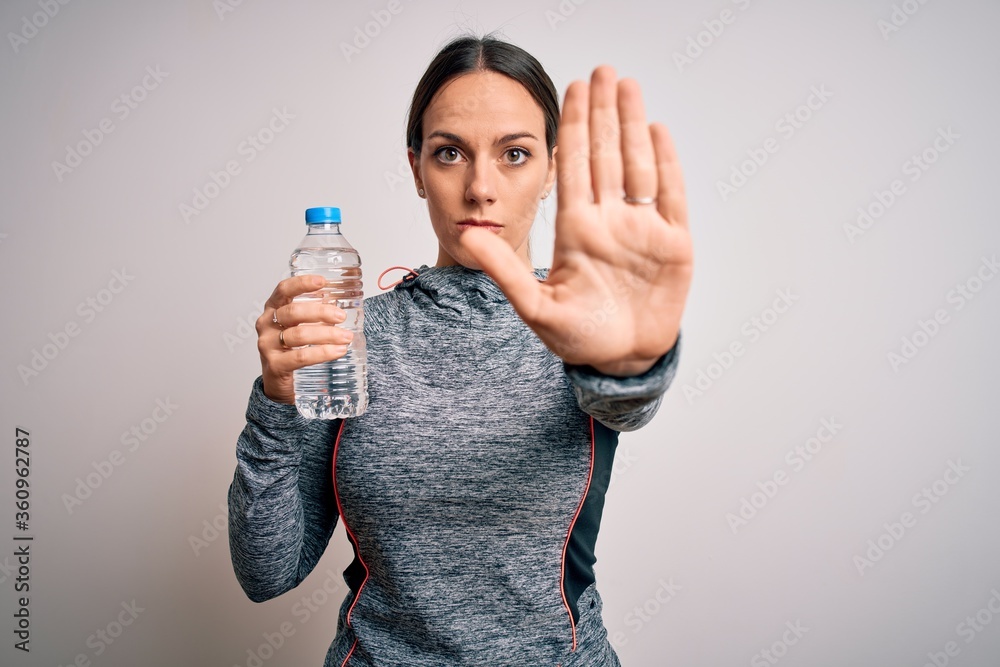 Young fitness woman wearing sport workout clothes drinking water from plastic bottle with open hand doing stop sign with serious and confident expression, defense gesture Young fitness woman wearing sport workout clothes drinking water from plastic bottle with open hand doing stop sign with serious and confident expression, defense gesture