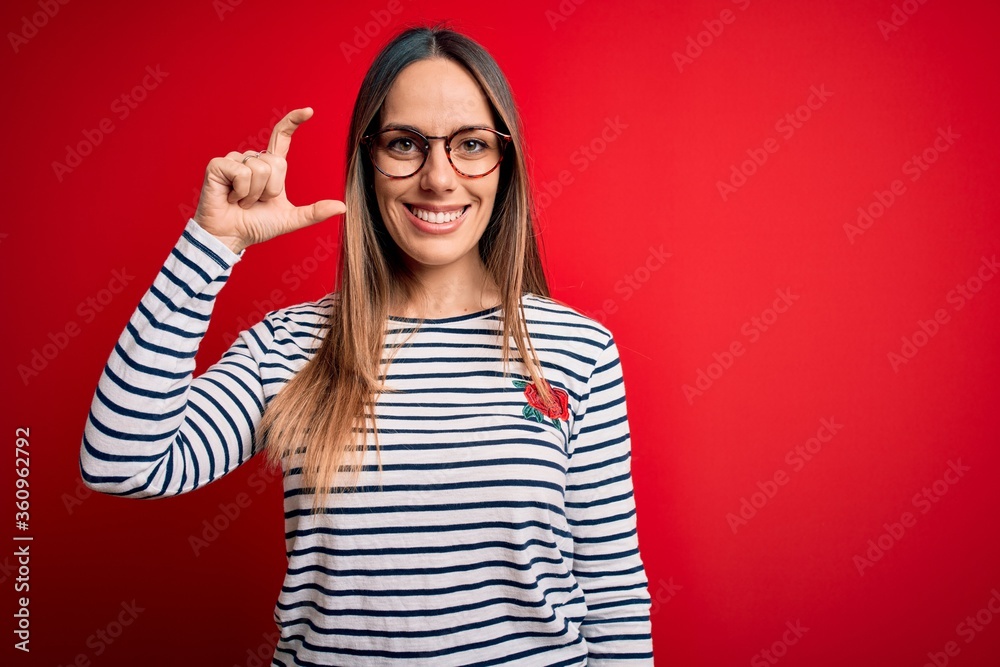 Young beautiful blonde woman with blue eyes wearing glasses standing over red background smiling and confident gesturing with hand doing small size sign with fingers looking and the camera. Measure