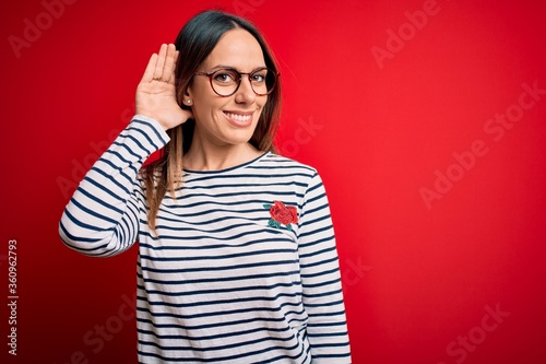 Young beautiful blonde woman with blue eyes wearing glasses standing over red background smiling with hand over ear listening an hearing to rumor or gossip. Deafness concept.