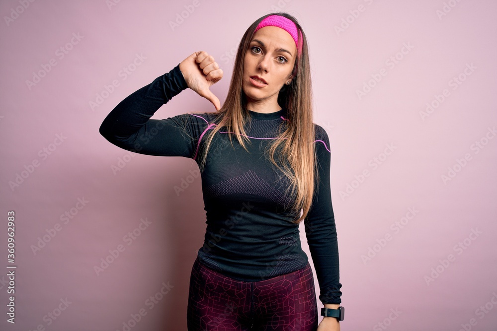 Young blonde fitness woman wearing sport workout clothes over isolated background looking unhappy and angry showing rejection and negative with thumbs down gesture. Bad expression. Young blonde fitness woman wearing sport workout clothes over isolated background looking unhappy and angry showing rejection and negative with thumbs down gesture. Bad expression.