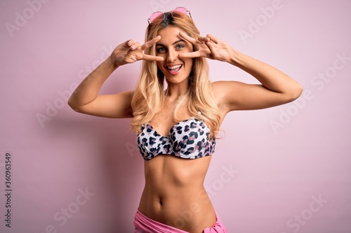 Young beautiful blonde woman on vacation wearing bikini over isolated pink background Doing peace symbol with fingers over face, smiling cheerful showing victory © Krakenimages.com