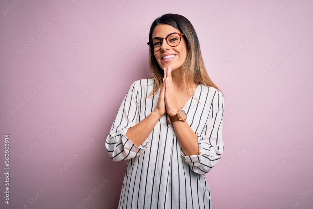 Young beautiful woman wearing casual striped t-shirt and glasses over pink background praying with hands together asking for forgiveness smiling confident. Young beautiful woman wearing casual striped t-shirt and glasses over pink background praying with hands together asking for forgiveness smiling confident.