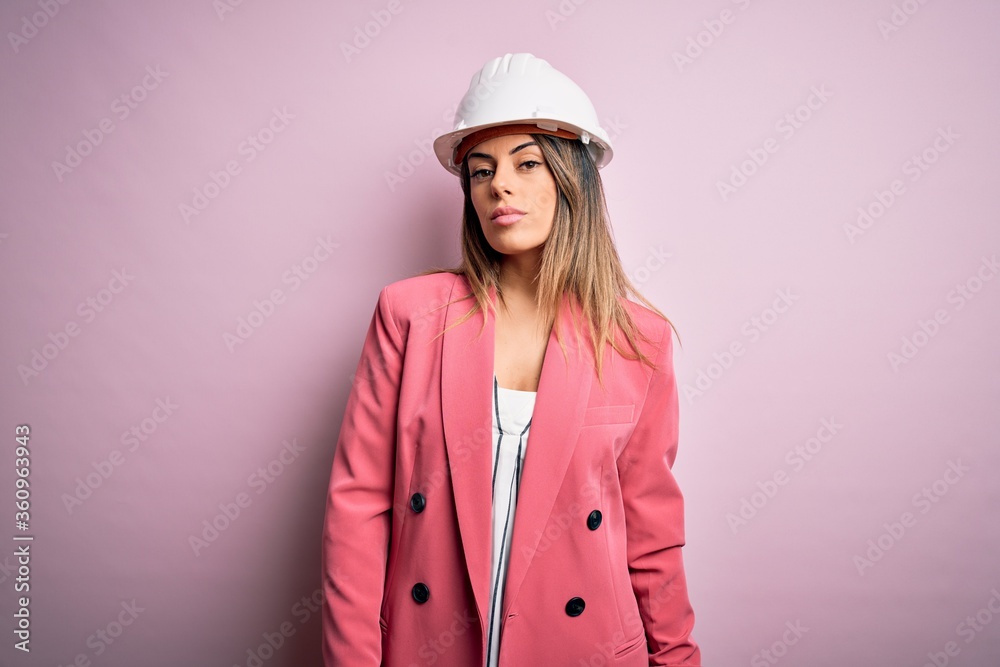 Young beautiful brunette architect woman wearing safety helmet over pink background Relaxed with serious expression on face. Simple and natural looking at the camera.