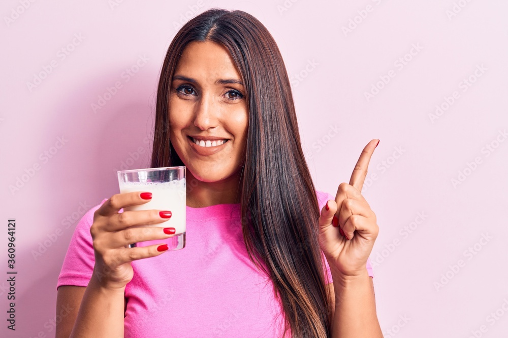 Young beautiful brunette woman drinking glass of milk smiling happy pointing with hand and finger to the side Young beautiful brunette woman drinking glass of milk smiling happy pointing with hand and finger to the side
