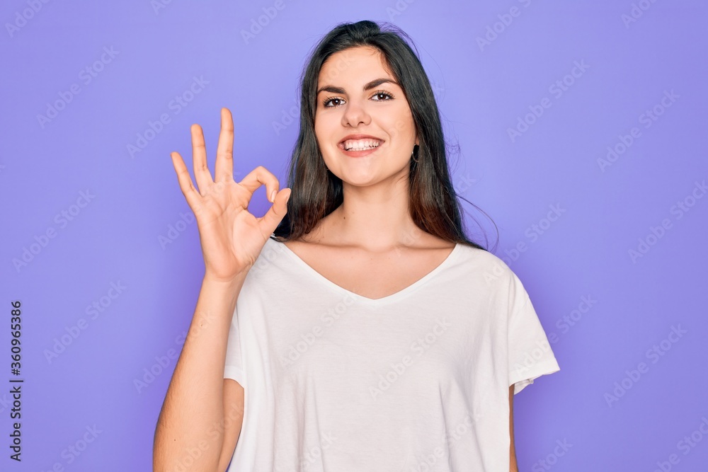 Young beautiful brunette woman wearing casual white t-shirt over purple background smiling positive doing ok sign with hand and fingers. Successful expression.