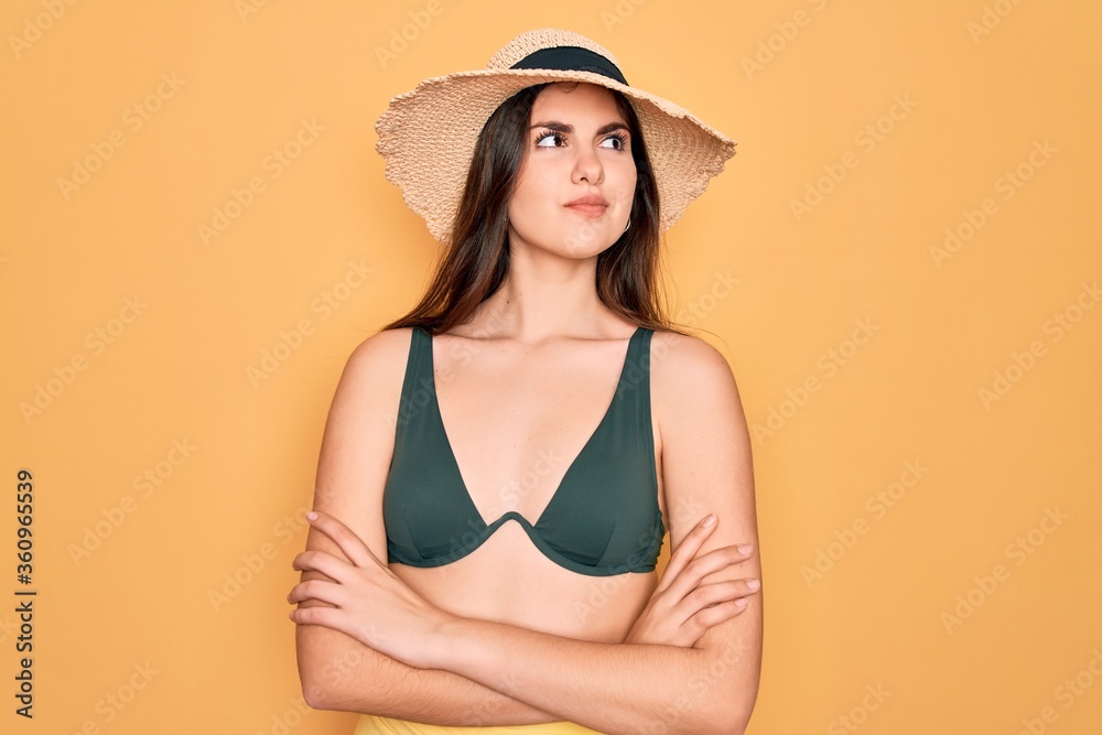 Young beautiful girl wearing swimwear bikini and summer sun hat over yellow background smiling looking to the side and staring away thinking. Young beautiful girl wearing swimwear bikini and summer sun hat over yellow background smiling looking to the side and staring away thinking.