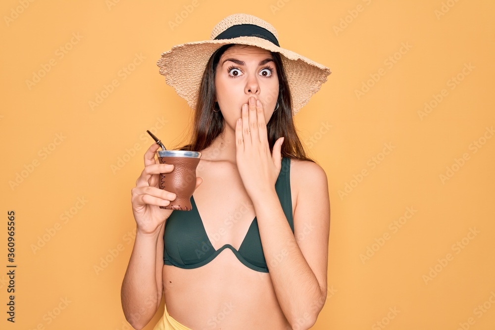 Young beautiful girl wearing swimwear bikini and summer hat drinking traditional mate drink cover mouth with hand shocked with shame for mistake, expression of fear, scared in silence, secret concept Young beautiful girl wearing swimwear bikini and summer hat drinking traditional mate drink cover mouth with hand shocked with shame for mistake, expression of fear, scared in silence, secret concept