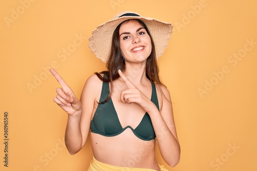 Young beautiful girl wearing swimwear bikini and summer sun hat over yellow background smiling and looking at the camera pointing with two hands and fingers to the side.