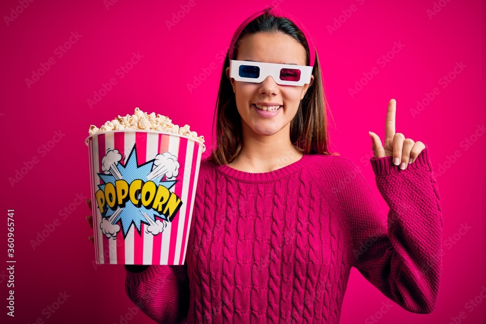 Obraz premium Young beautiful girl watching movie using 3d glasses eating box with popcorns with a big smile on face, pointing with hand finger to the side looking at the camera.