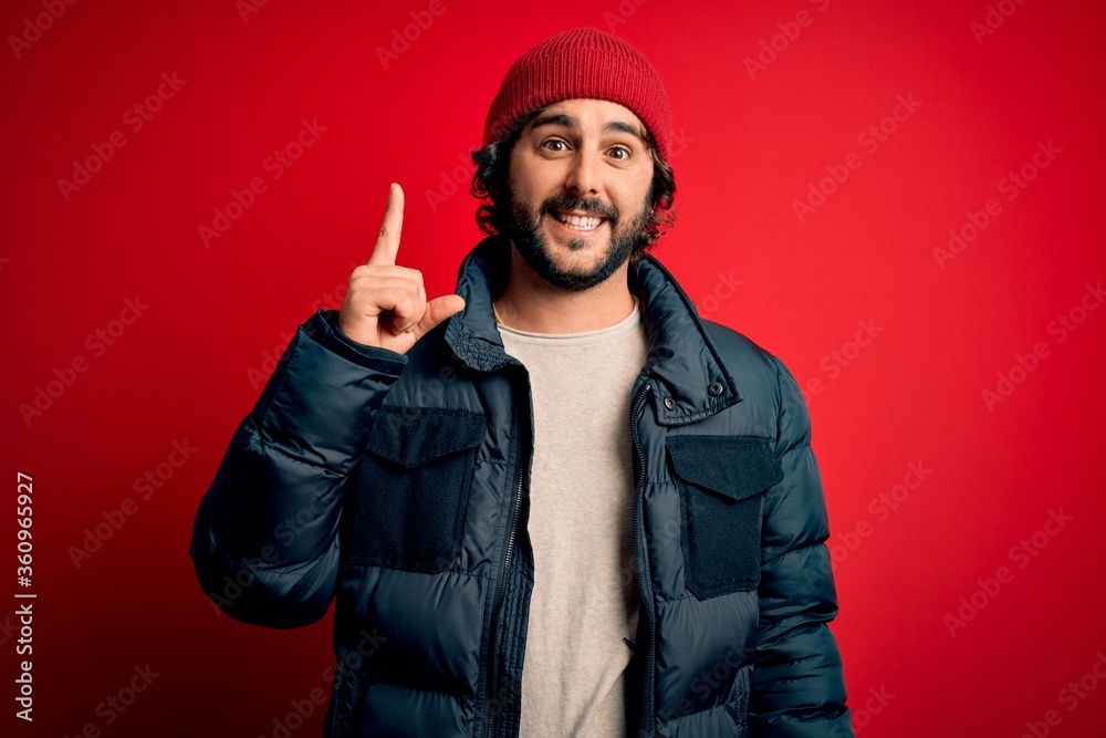 Young handsome man with beard wearing casual coat and wool cap over red background pointing finger up with successful idea. Exited and happy. Number one. Young handsome man with beard wearing casual coat and wool cap over red background pointing finger up with successful idea. Exited and happy. Number one.