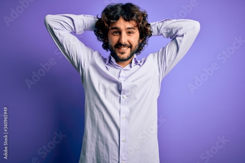 Young handsome business man with beard wearing shirt standing over purple background relaxing and stretching, arms and hands behind head and neck smiling happy © Krakenimages.com