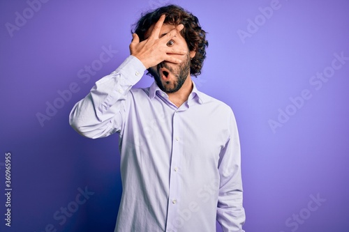 Young handsome business man with beard wearing shirt standing over purple background peeking in shock covering face and eyes with hand, looking through fingers with embarrassed expression. © Krakenimages.com