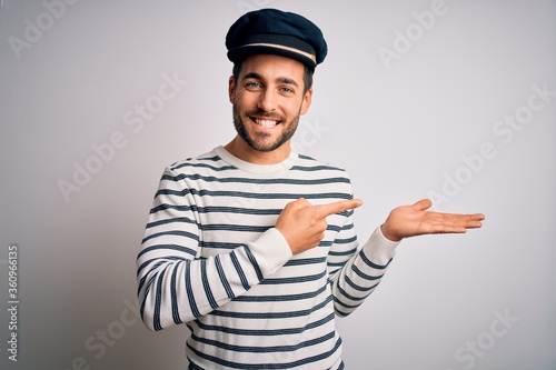 Young handsome sailor man with beard wearing navy striped uniform and captain hat amazed and smiling to the camera while presenting with hand and pointing with finger. photo