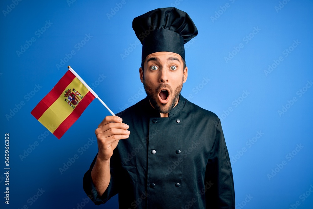 Young handsome cooker man with beard wearing uniform holding spain spanish flag scared in shock with a surprise face, afraid and excited with fear expression Young handsome cooker man with beard wearing uniform holding spain spanish flag scared in shock with a surprise face, afraid and excited with fear expression