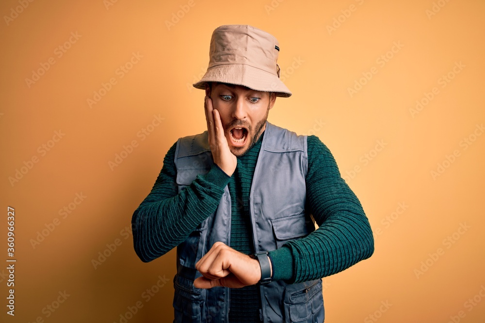 Handsome tourist man with beard on vacation wearing explorer hat over yellow background Looking at the watch time worried, afraid of getting late Handsome tourist man with beard on vacation wearing explorer hat over yellow background Looking at the watch time worried, afraid of getting late