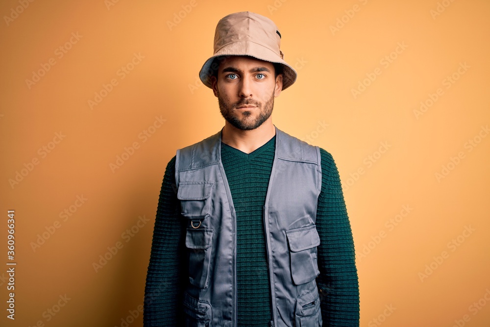 Handsome tourist man with beard on vacation wearing explorer hat over yellow background Relaxed with serious expression on face. Simple and natural looking at the camera. Handsome tourist man with beard on vacation wearing explorer hat over yellow background Relaxed with serious expression on face. Simple and natural looking at the camera.