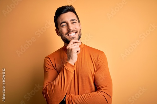 Young handsome man with beard wearing casual sweater standing over yellow background looking confident at the camera with smile with crossed arms and hand raised on chin. Thinking positive.
