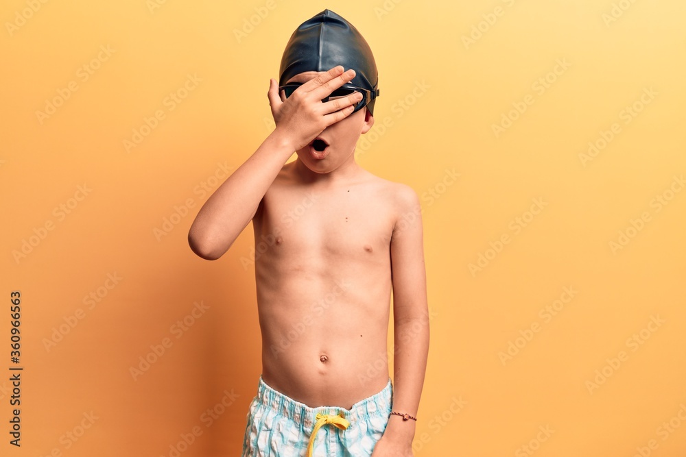 Cute blond kid wearing swimwear and swimmer glasses peeking in shock covering face and eyes with hand, looking through fingers afraid