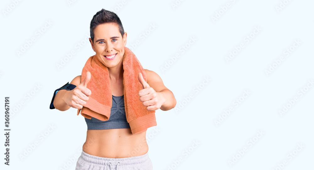 Young woman with short hair wearing sportswear and towel using smartphone approving doing positive gesture with hand, thumbs up smiling and happy for success. winner gesture. Young woman with short hair wearing sportswear and towel using smartphone approving doing positive gesture with hand, thumbs up smiling and happy for success. winner gesture.