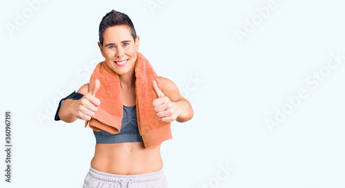 Young woman with short hair wearing sportswear and towel using smartphone approving doing positive gesture with hand, thumbs up smiling and happy for success. winner gesture.