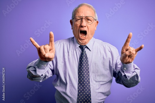 Grey haired senior business man wearing glasses standing over purple isolated background shouting with crazy expression doing rock symbol with hands up. Music star. Heavy concept.