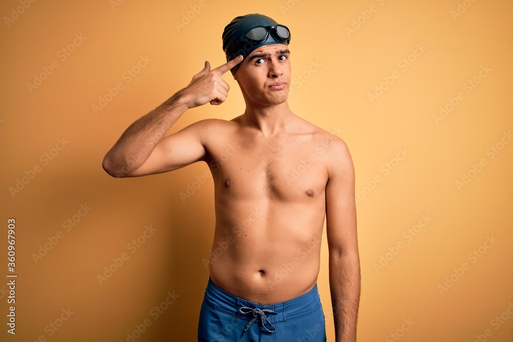 Young handsome man shirtless wearing swimsuit and swim cap over isolated yellow background pointing unhappy to pimple on forehead, ugly infection of blackhead. Acne and skin problem