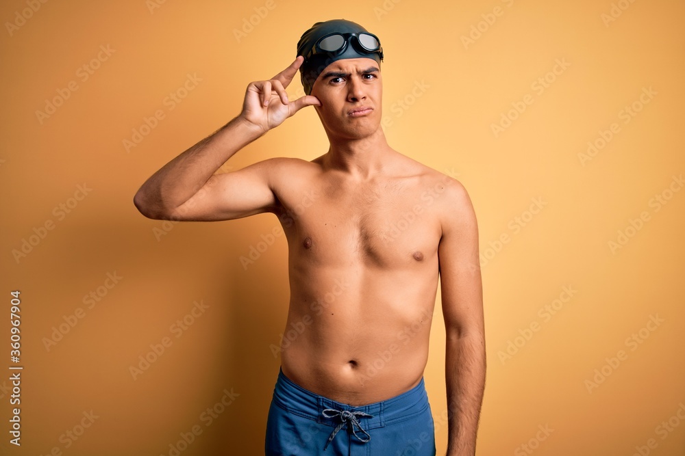 Young handsome man shirtless wearing swimsuit and swim cap over isolated yellow background worried and stressed about a problem with hand on forehead, nervous and anxious for crisis