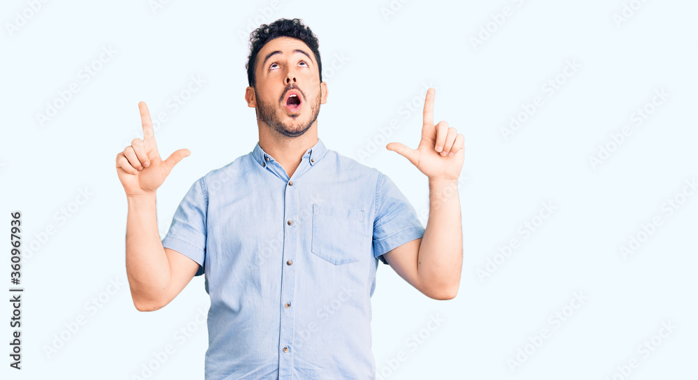 Young hispanic man wearing casual clothes amazed and surprised looking up and pointing with fingers and raised arms. Young hispanic man wearing casual clothes amazed and surprised looking up and pointing with fingers and raised arms.