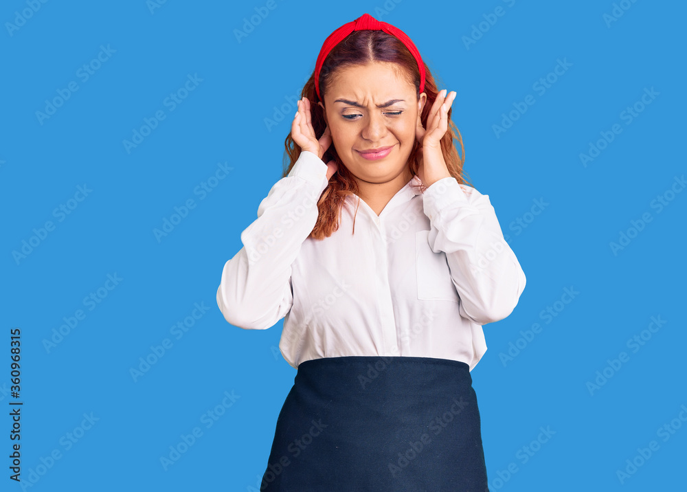 Young latin woman wearing waitress apron covering ears with fingers with annoyed expression for the noise of loud music. deaf concept.