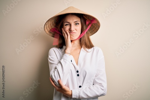 Young beautiful redhead woman wearing asian traditional conical hat over white background thinking looking tired and bored with depression problems with crossed arms.