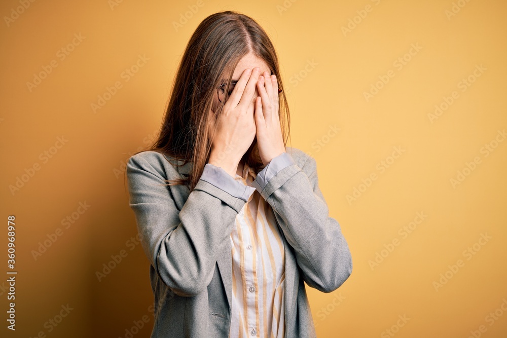 Young beautiful redhead woman wearing jacket and glasses over isolated yellow background with sad expression covering face with hands while crying. Depression concept. Young beautiful redhead woman wearing jacket and glasses over isolated yellow background with sad expression covering face with hands while crying. Depression concept.