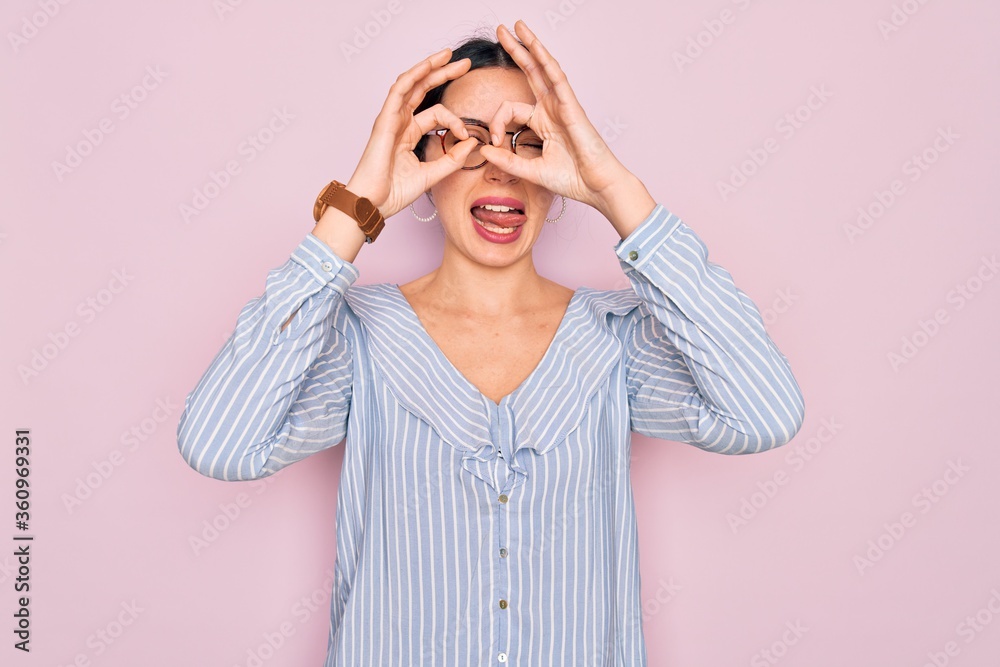 Young beautiful woman wearing casual striped shirt and glasses over pink background doing ok gesture like binoculars sticking tongue out, eyes looking through fingers. Crazy expression. Young beautiful woman wearing casual striped shirt and glasses over pink background doing ok gesture like binoculars sticking tongue out, eyes looking through fingers. Crazy expression.