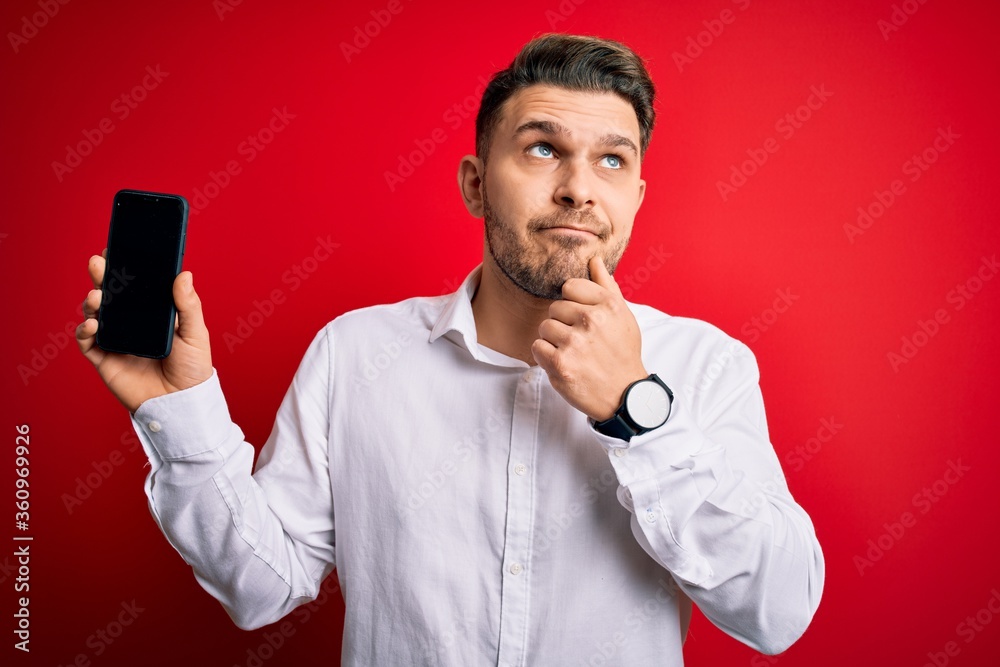 Young business man with blue eyes showing smartphone screen over red background serious face thinking about question, very confused idea Young business man with blue eyes showing smartphone screen over red background serious face thinking about question, very confused idea