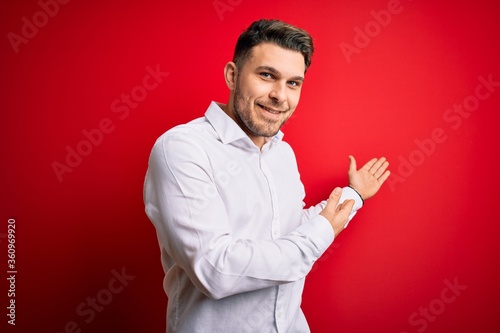 Young business man with blue eyes wearing elegant shirt standing over red isolated background Inviting to enter smiling natural with open hand