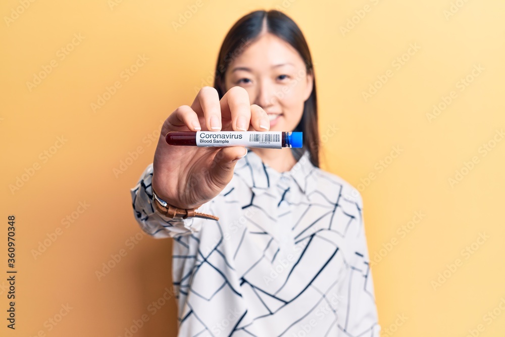 Young beautiful chinese woman holding test tube with coronavirus blood sample looking positive and happy standing and smiling with a confident smile showing teeth Young beautiful chinese woman holding test tube with coronavirus blood sample looking positive and happy standing and smiling with a confident smile showing teeth