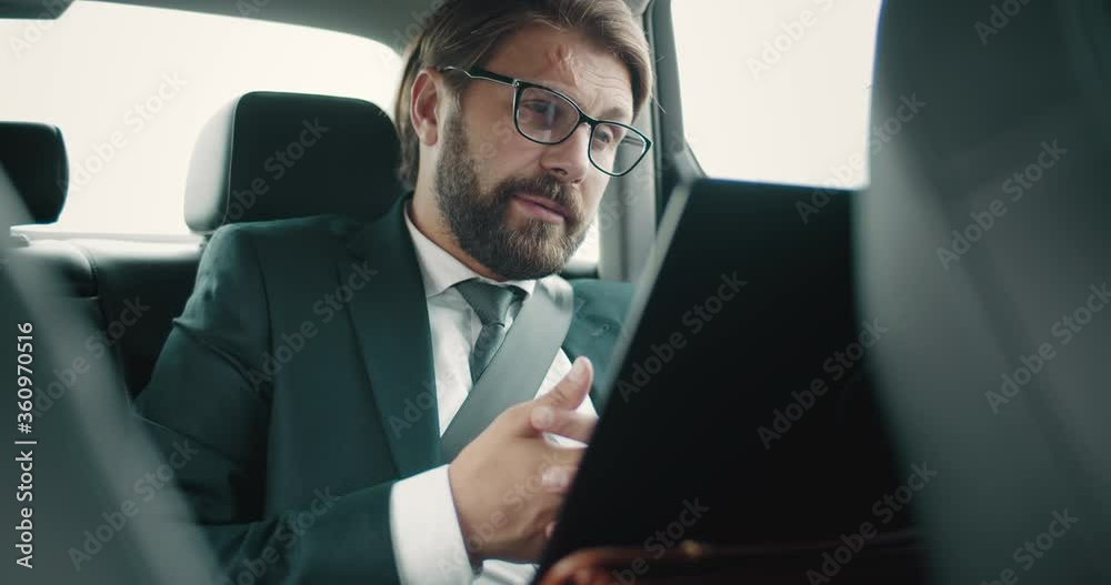 Mature serious man in eyeglasses talking with business partners through video call on portable computer. Bearded businessman in formal outfit using laptop for conversation in auto. Mature serious man in eyeglasses talking with business partners through video call on portable computer. Bearded businessman in formal outfit using laptop for conversation in auto.